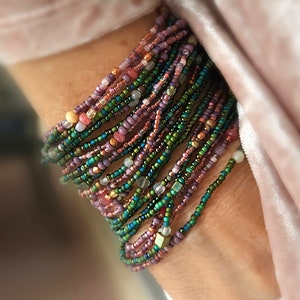 May include: A stack of colorful beaded bracelets in shades of green, purple, pink, and gold. The bracelets are made of small, round beads and are layered on top of each other.