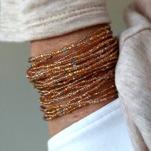 Könnte beinhalten: Ein mehrreihiges Armband aus kleinen braunen, goldenen und durchsichtigen Perlen. Das Armband ist um das Handgelenk einer Person gewickelt.