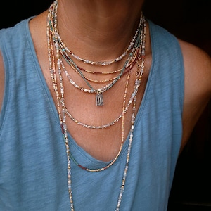 May include: A multi-strand necklace set featuring gold, red, teal, and clear beads. A small silver pendant is suspended from one strand. The necklaces are worn over a blue vest top.