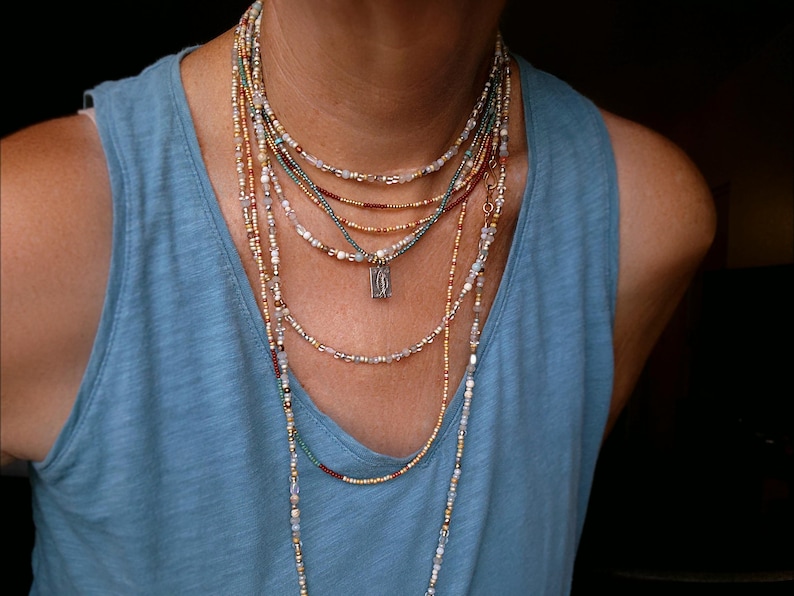 May include: A multi-strand necklace set featuring gold, red, teal, and clear beads. A small silver pendant is suspended from one strand. The necklaces are worn over a blue vest top.