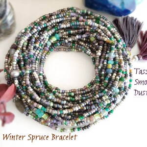 May include: A multi-strand bracelet made of small, colorful beads in shades of green, gray, silver, and purple. The bracelet has a pearl accent and two tassels, one in a dusty plum color and the other in a smoke color. The bracelet is called "Winter Spruce Bracelet".
