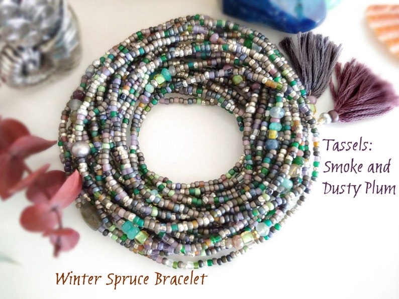 May include: A multi-strand bracelet made of small, colorful beads in shades of green, gray, silver, and purple. The bracelet has a pearl accent and two tassels, one in a dusty plum color and the other in a smoke color. The bracelet is called "Winter Spruce Bracelet".