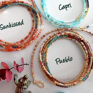 May include: Three beaded necklaces in the colours "Sunkissed", "Capri", and "Pueblo" are displayed. "Sunkissed" features orange and red beads. "Capri" has blue and white beads. "Pueblo" has brown, tan, and teal beads. Each necklace is coiled.
