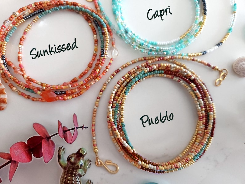 May include: Three beaded necklaces in the colours "Sunkissed", "Capri", and "Pueblo" are displayed. "Sunkissed" features orange and red beads. "Capri" has blue and white beads. "Pueblo" has brown, tan, and teal beads. Each necklace is coiled.