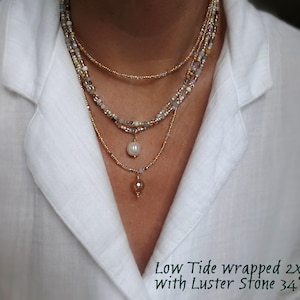 May include: A multi-strand necklace with gold, white, and neutral-toned beads. It includes a pearl pendant and a small, amber-coloured stone pendant. The text "Low Tide wrapped 2x With Luster Stone 34" is visible, with a total length of 86 cm.