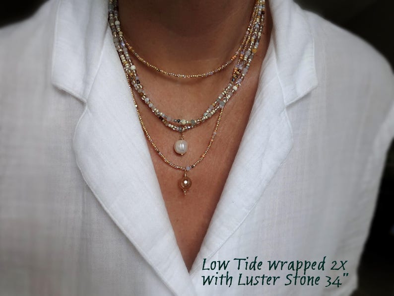 May include: A multi-strand necklace with gold, white, and neutral-toned beads. It includes a pearl pendant and a small, amber-coloured stone pendant. The text "Low Tide wrapped 2x With Luster Stone 34" is visible, with a total length of 86 cm.