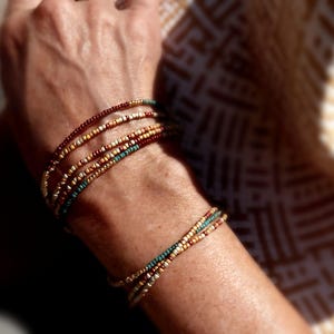 May include: Beaded bracelets in brown, gold, and teal. The bracelets are made of small, colourful beads, creating a layered effect. The bracelets are worn on the wrist. The image shows the bracelets on a person's arm.