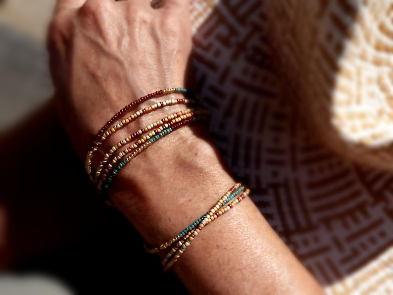 May include: Beaded bracelets in brown, gold, and teal. The bracelets are made of small, colourful beads, creating a layered effect. The bracelets are worn on the wrist. The image shows the bracelets on a person's arm.