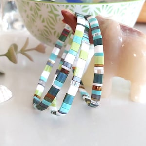 May include: Stackable bracelets with a mix of colors including teal, white, green, and brown. The bracelets are made of small, flat beads and are arranged in a stack. The bracelets are on a white surface with decorative items in the background.