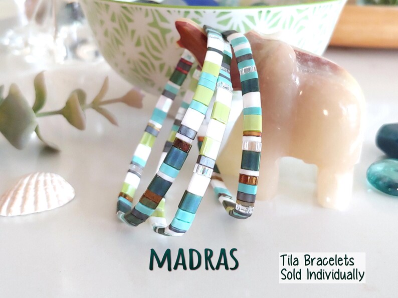 Peut inclure: Trois bracelets compos&eacute;s de perles carr&eacute;es et plates multicolores dans des tons de bleu sarcelle, vert lime, blanc et bronze m&eacute;tallique. Le texte MADRAS et Tila Bracelets Sold Individually est imprim&eacute; en vert fonc&eacute; sous les bracelets.