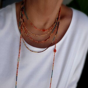 May include: A multi-strand beaded necklace in shades of orange, red, gold, and teal. The necklace features small beads and a long, dangling strand. The necklace is worn over a white top.