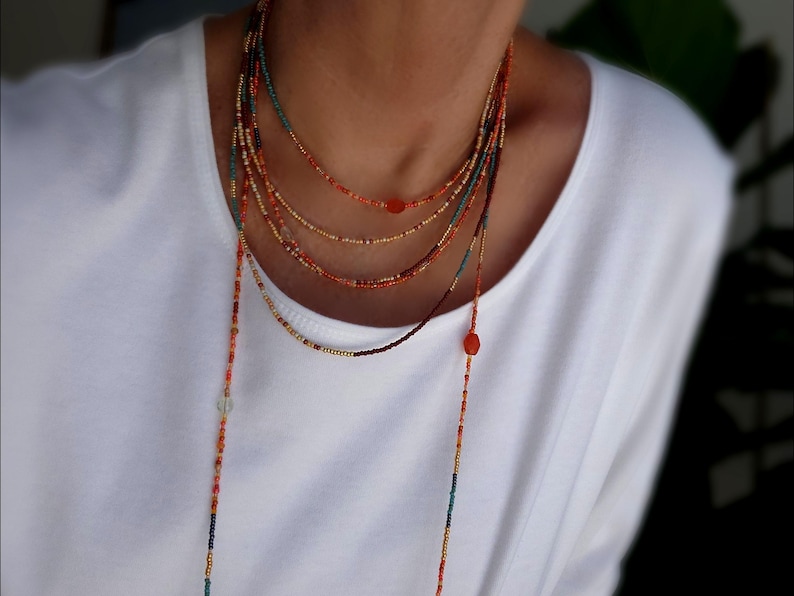 May include: A multi-strand beaded necklace in shades of orange, red, gold, and teal. The necklace features small beads and a long, dangling strand. The necklace is worn over a white top.