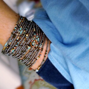 May include: A close-up of a person's wrist wearing a stack of beaded bracelets. The bracelets are made of small, gray, brown, and turquoise beads. The bracelets are layered on top of each other, creating a thick, textured look.