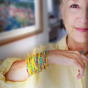 K&ouml;nnte beinhalten: Eine Frau tr&auml;gt ein gelbes Hemd mit einem Stapel bunter Perlenarmb&auml;nder am Handgelenk. Die Armb&auml;nder bestehen aus kleinen, runden Perlen in den Farben Gelb, Blau, T&uuml;rkis und Rot.