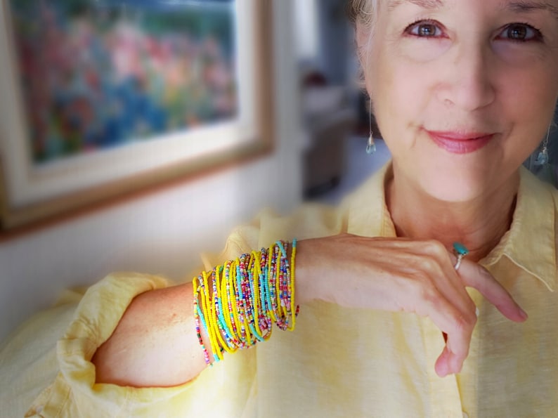 K&ouml;nnte beinhalten: Eine Frau tr&auml;gt ein gelbes Hemd mit einem Stapel bunter Perlenarmb&auml;nder am Handgelenk. Die Armb&auml;nder bestehen aus kleinen, runden Perlen in den Farben Gelb, Blau, T&uuml;rkis und Rot.