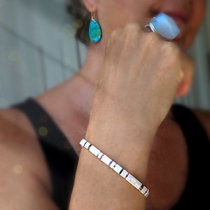 Peut inclure: Un bracelet compos&eacute; de tuiles rectangulaires blanches aux reflets iris&eacute;s, une boucle d'oreille turquoise en forme de goutte avec un fil d'or, et une bague orn&eacute;e d'une grosse pierre bleue translucide facett&eacute;e sur un anneau en fil d'argent.