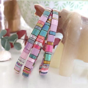 May include: Three thin bracelets composed of small, square, multi-coloured tiles in shades of pink, teal, grey, and metallic gold. The tiles are arranged in a repeating pattern, creating a vibrant, textured look. The bracelets are stacked together.