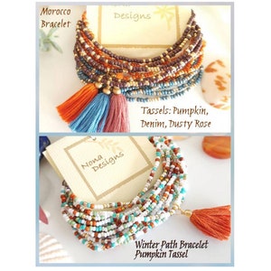 May include: Two multi-strand beaded bracelets with tassels. The top bracelet is brown, orange, and blue with three tassels. The bottom bracelet is white, orange, and turquoise with one tassel. The bracelets are labeled "Morocco Bracelet" and "Winter Path Bracelet Pumpkin Tassel".