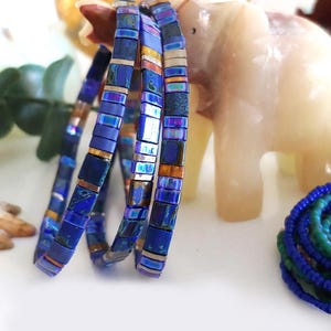 May include: Three blue beaded bracelets with iridescent and gold accents. The bracelets are made of small, rectangular beads and are coiled. A blue and green beaded bracelet is in the background, along with a white elephant statue.