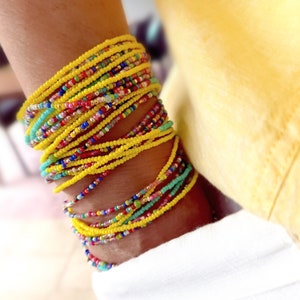 K&ouml;nnte beinhalten: Ein Stapel bunter Perlenarmb&auml;nder, haupts&auml;chlich gelb, mit einigen t&uuml;rkisfarbenen, roten und rosafarbenen Perlen. Die Armb&auml;nder werden am Handgelenk getragen.