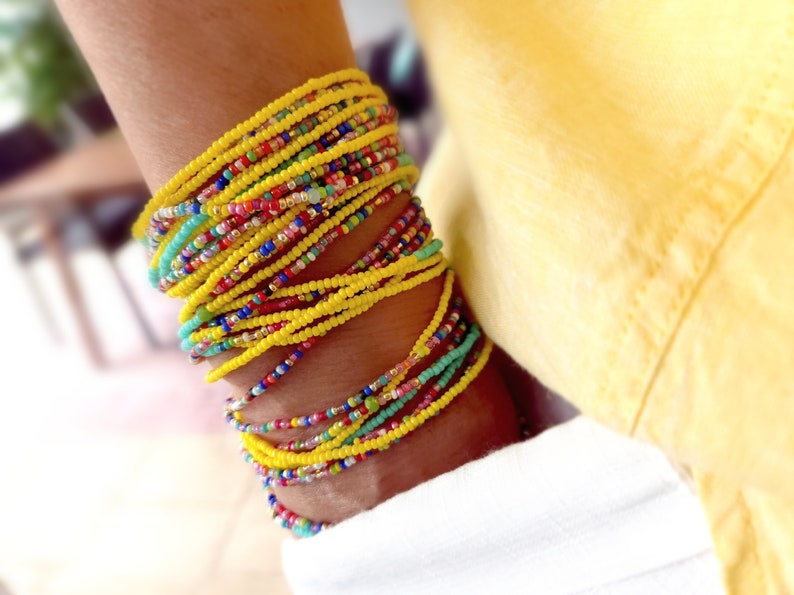 K&ouml;nnte beinhalten: Ein Stapel bunter Perlenarmb&auml;nder, haupts&auml;chlich gelb, mit einigen t&uuml;rkisfarbenen, roten und rosafarbenen Perlen. Die Armb&auml;nder werden am Handgelenk getragen.