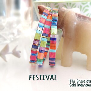 Peut inclure: Trois bracelets Tila empil&eacute;s compos&eacute;s de perles de verre carr&eacute;es multicolores dans des tons de rose, orange, bleu, vert et or m&eacute;tallique. Le texte FESTIVAL et Tila Bracelets Sold Individually est imprim&eacute; en noir au bas de l'image.