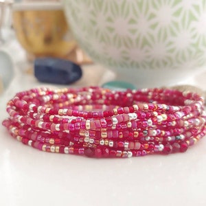 May include: A stack of beaded bracelets in shades of red, pink, and gold. The bracelets are made of small, round beads and are layered together. The bracelets are likely handmade and are a fashion accessory.