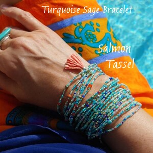 May include: Turquoise Sage Bracelet with a salmon tassel. The bracelet is made of multiple strands of tiny beads in shades of turquoise, blue, and gold. The tassel is a bright orange color.