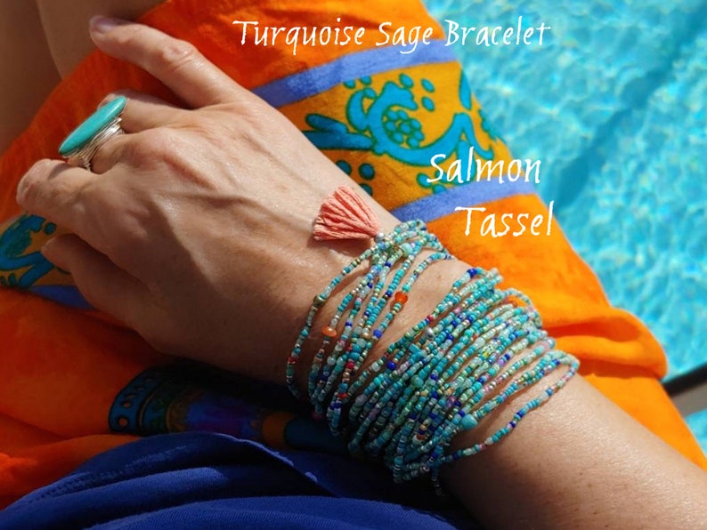 May include: Turquoise Sage Bracelet with a salmon tassel. The bracelet is made of multiple strands of tiny beads in shades of turquoise, blue, and gold. The tassel is a bright orange color.