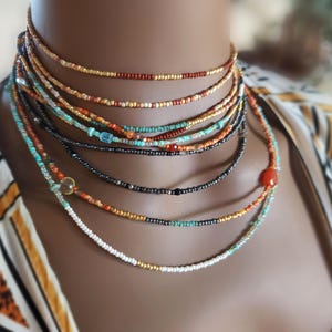 May include: A selection of layered beaded necklaces in shades of red, orange, turquoise, and black. The necklaces are composed of tiny beads and are worn around the neck. The background is blurred, with a patterned top visible.