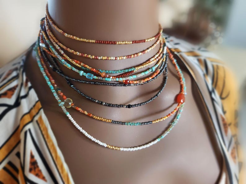 May include: A selection of layered beaded necklaces in shades of red, orange, turquoise, and black. The necklaces are composed of tiny beads and are worn around the neck. The background is blurred, with a patterned top visible.