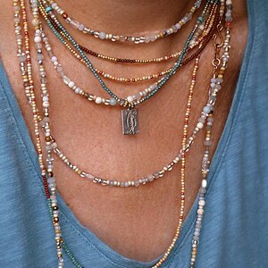 May include: A multi-strand beaded necklace in shades of gold, teal, and white. A small, rectangular pendant with a fish design is visible. The necklaces are worn over a blue top. The beads are of varying sizes and materials.