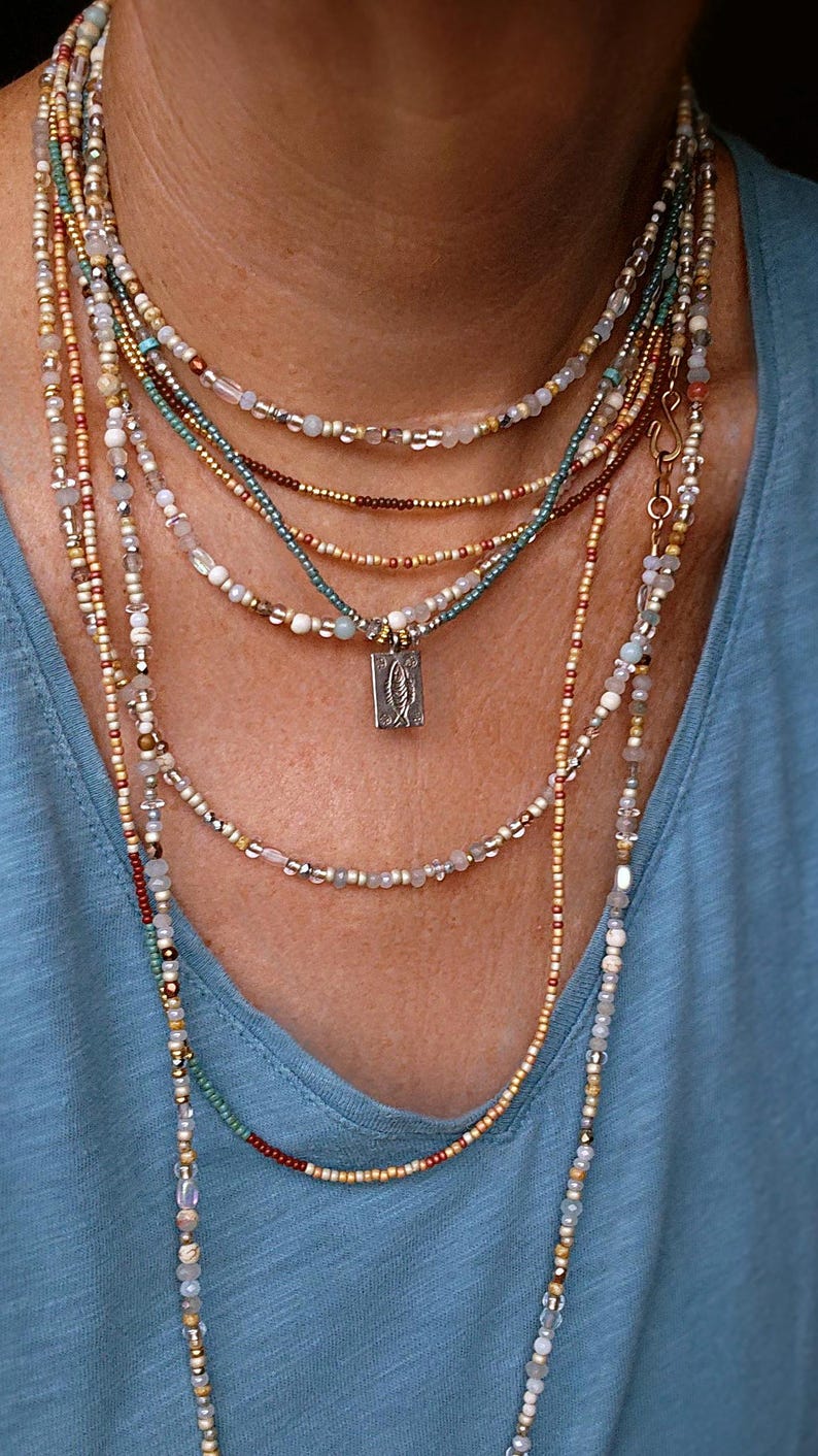 May include: A multi-strand beaded necklace in shades of gold, teal, and white. A small, rectangular pendant with a fish design is visible. The necklaces are worn over a blue top. The beads are of varying sizes and materials.