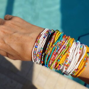 May include: A wrist displaying a collection of colourful beaded bracelets. The bracelets feature a variety of hues, such as white, yellow, black, and turquoise. The small beads create a layered effect. A silver ring with a light blue stone is also visible.