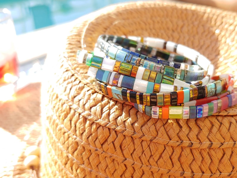 Peut inclure: Une pile de bracelets de perles multicolores compos&eacute;s de perles plates et rectangulaires dans des tons de blanc, or m&eacute;tallique, bleu iris&eacute;, vert, orange, noir et rose. Les bracelets sont dispos&eacute;s en cercle avec une finition brillante.