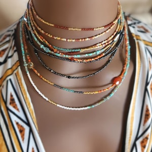 May include: A multi-strand beaded necklace set in shades of gold, brown, black, turquoise, and orange. The necklaces are displayed against a patterned yellow and white top. The beads are small and delicate.