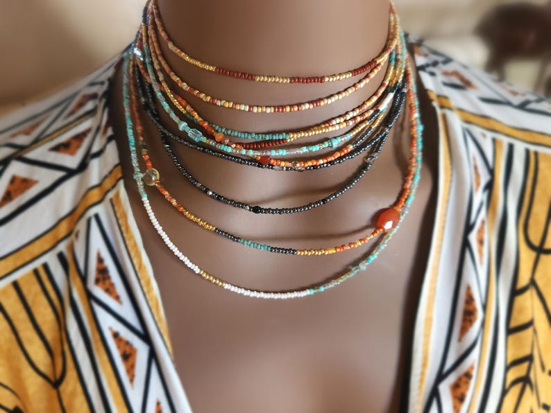 May include: A multi-strand beaded necklace set in shades of gold, brown, black, turquoise, and orange. The necklaces are displayed against a patterned yellow and white top. The beads are small and delicate.