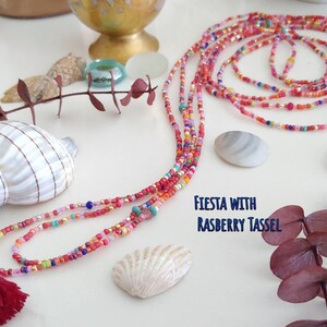 May include: Multicolored beaded necklace with a red tassel. The necklace is made of small, colorful beads and has a long, flowing design. The tassel is made of red yarn and is attached to the end of the necklace. The text "FIESTA WITH RASPBERRY TASSEL" is written on the white surface below the necklace.
