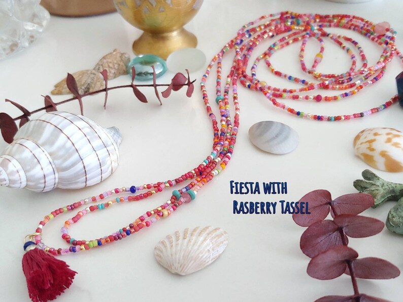 May include: Multicolored beaded necklace with a red tassel. The necklace is made of small, colorful beads and has a long, flowing design. The tassel is made of red yarn and is attached to the end of the necklace. The text "FIESTA WITH RASPBERRY TASSEL" is written on the white surface below the necklace.