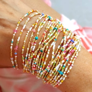 K&ouml;nnte beinhalten: Ein Stapel bunter Perlenarmb&auml;nder in Wei&szlig;, Gold und verschiedenen leuchtenden Farben. Die Armb&auml;nder sind &uuml;bereinander am Handgelenk einer Person gelegt.