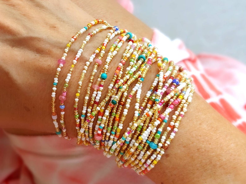 K&ouml;nnte beinhalten: Ein Stapel bunter Perlenarmb&auml;nder in Wei&szlig;, Gold und verschiedenen leuchtenden Farben. Die Armb&auml;nder sind &uuml;bereinander am Handgelenk einer Person gelegt.