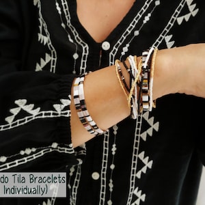 Peut inclure: Une collection de bracelets Tuxedo Tila compos&eacute;e de perles noires, blanches et dor&eacute;es m&eacute;tallis&eacute;es. Les bracelets sont dispos&eacute;s en deux groupes distincts sur le poignet, mettant en valeur des perles carr&eacute;es Tila et des perles rondes plus petites.