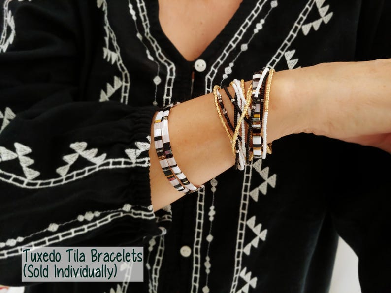 Peut inclure: Une collection de bracelets Tuxedo Tila compos&eacute;e de perles noires, blanches et dor&eacute;es m&eacute;tallis&eacute;es. Les bracelets sont dispos&eacute;s en deux groupes distincts sur le poignet, mettant en valeur des perles carr&eacute;es Tila et des perles rondes plus petites.