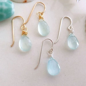 May include: Four pairs of aquamarine teardrop earrings. Two pairs have gold-colored ear wires, and two have silver-colored ear wires. The aquamarine stones are faceted and have a pale blue color. The earrings are displayed on a white surface.
