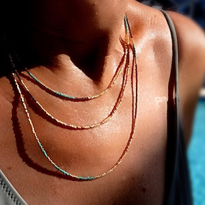 May include: A multi-strand necklace with small beads in gold, turquoise, and brown. The necklace is composed of three delicate strands. The background is blurred, with a person's skin visible. The necklace is a piece of jewellery.