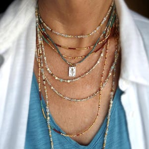 May include: A multi-strand necklace set with beads in gold, teal, and red. A small pendant hangs from one strand. The necklaces are worn over a blue top and a white collared shirt. The image shows a close-up of the neck and upper chest.