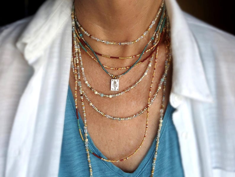 May include: A multi-strand necklace set with beads in gold, teal, and red. A small pendant hangs from one strand. The necklaces are worn over a blue top and a white collared shirt. The image shows a close-up of the neck and upper chest.