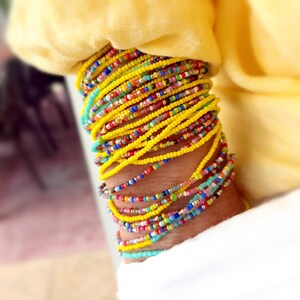 K&ouml;nnte beinhalten: Ein Stapel bunter Perlenarmb&auml;nder, haupts&auml;chlich gelb, mit einigen t&uuml;rkisfarbenen, rosa und roten Perlen. Die Armb&auml;nder werden am Handgelenk getragen.