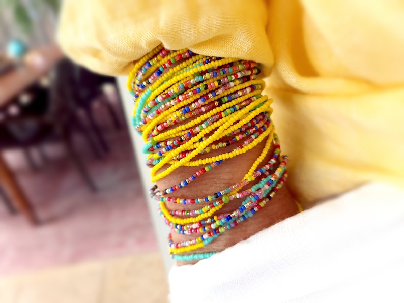 K&ouml;nnte beinhalten: Ein Stapel bunter Perlenarmb&auml;nder, haupts&auml;chlich gelb, mit einigen t&uuml;rkisfarbenen, rosa und roten Perlen. Die Armb&auml;nder werden am Handgelenk getragen.