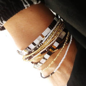 May include: A stack of multiple bracelets featuring a variety of beads, including flat square black and white tiles, metallic gold spheres, small clear glass beads, and thin strands of tiny black and white seed beads arranged in a layered design.